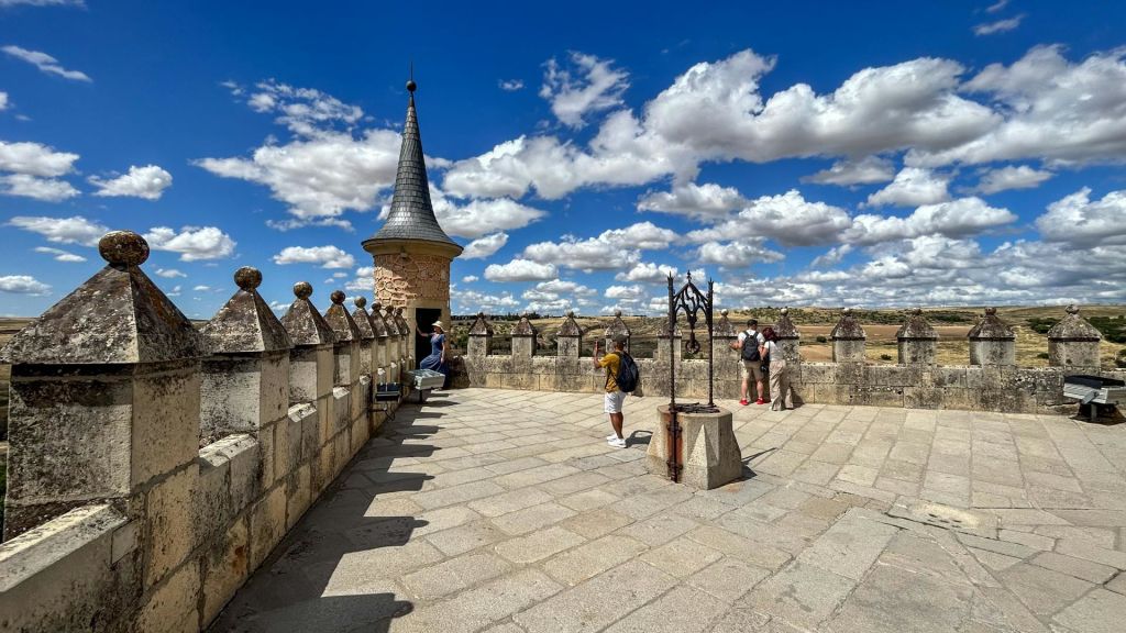 구름이 깔린 푸른 하늘 아래, 돌로 장식된 성곽 테라스, 원뿔형 타워, 철제 우물 구조물이 있는 사람들.