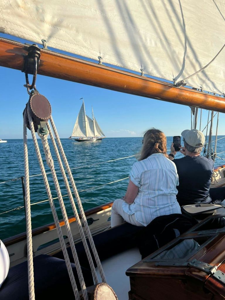 요트를 탄 두 사람이 맑고 푸른 하늘 아래 잔잔한 바다에서 먼 스쿠너를 관찰하고 사진을 찍고 있습니다.