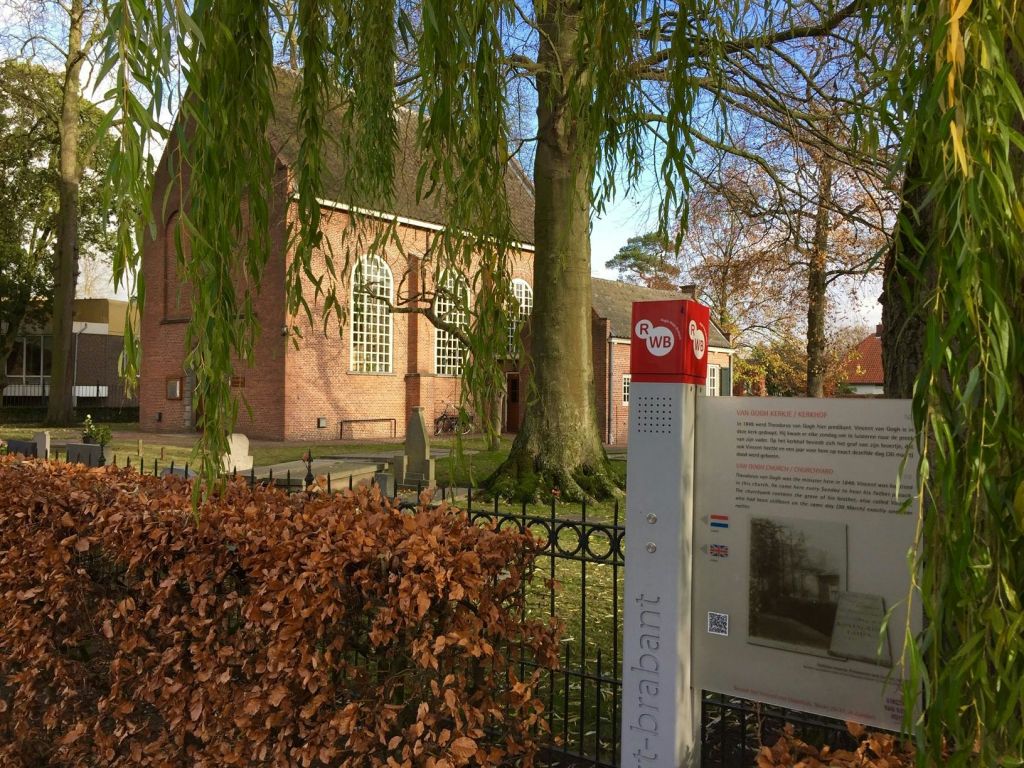 나무 뒤에 아치형 창문이 있고 울타리가 있는 벽돌 건물. 정보 표지판과 전경의 울타리.