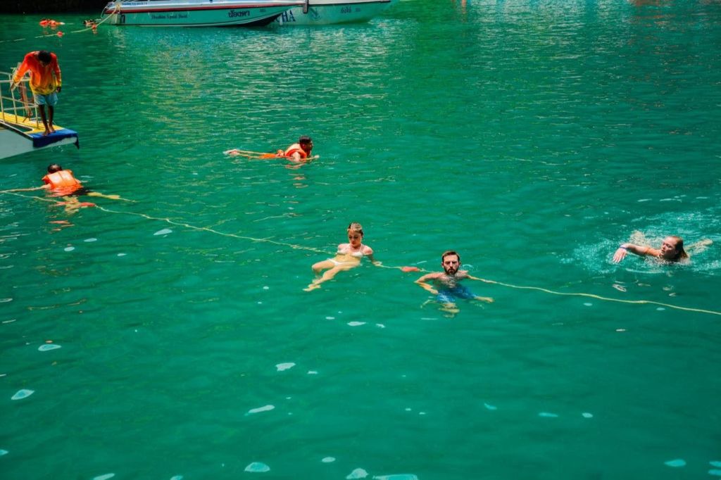 보트 근처의 맑고 푸른 물에서 수영하는 사람들이 있으며, 일부는 떠다니는 밧줄을 잡고 있습니다.