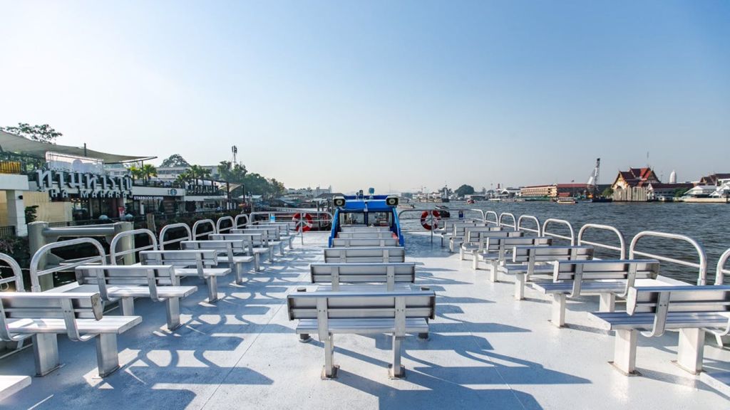 나룻배의 상부 갑판에는 벤치가 줄지어 있고, 측면에는 구명 부이가 있으며, 잔잔한 강에는 건물과 나무가 배경으로 있습니다.