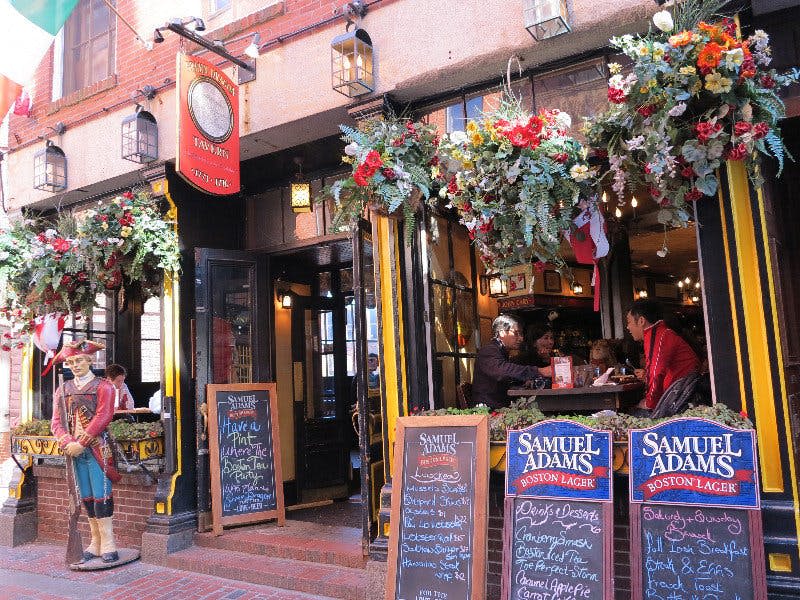 꽃 장식, 칠판 메뉴, 후원자가 앉아 있는 활기찬 펍 입구. Samuel Adams 광고가 눈에 띕니다.