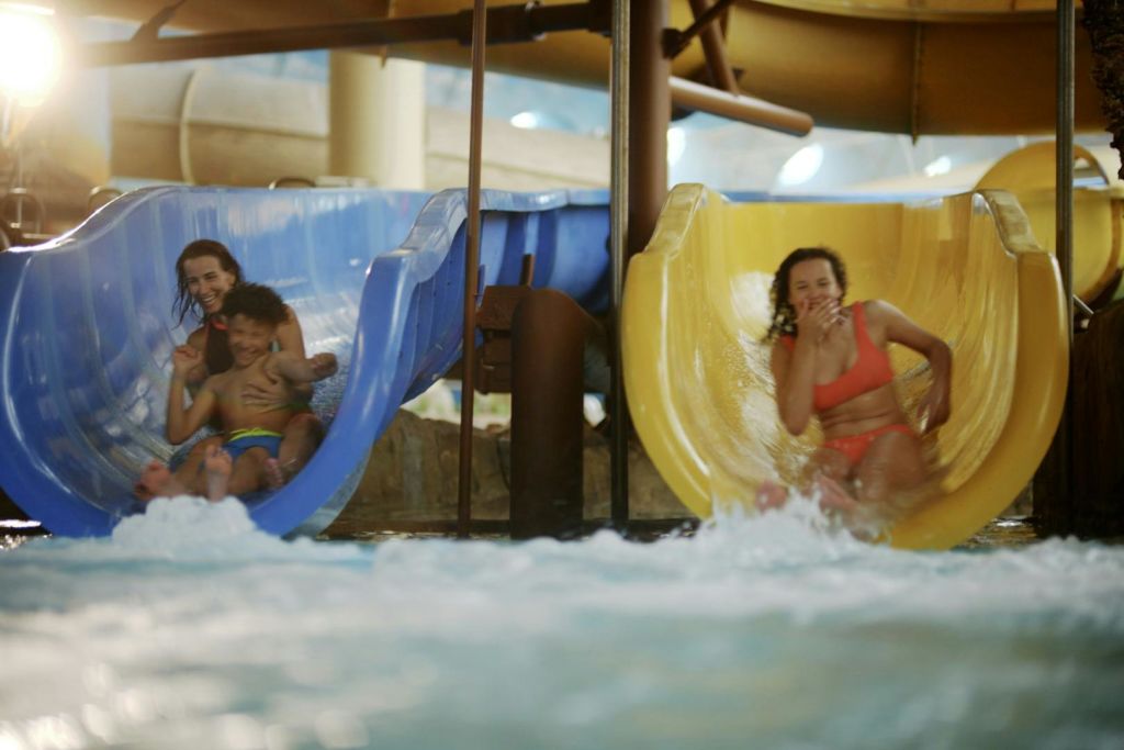 한 여성과 어린이가 푸른 물 미끄럼틀을 타고 실내 워터파크의 수영장으로 미끄러져 들어오고 있습니다.