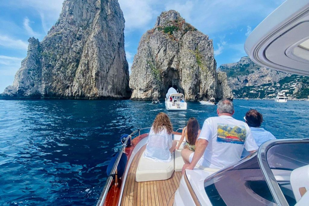 청명한 하늘 아래 푸른 물 위로 천연 아치가 있는 커다란 바위 절벽에 접근하는 보트를 탄 사람들.