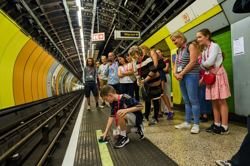 한 무리의 사람들이 지하철 플랫폼에서 지상의 작은 물체와 상호 작용하는 소년을 관찰합니다.