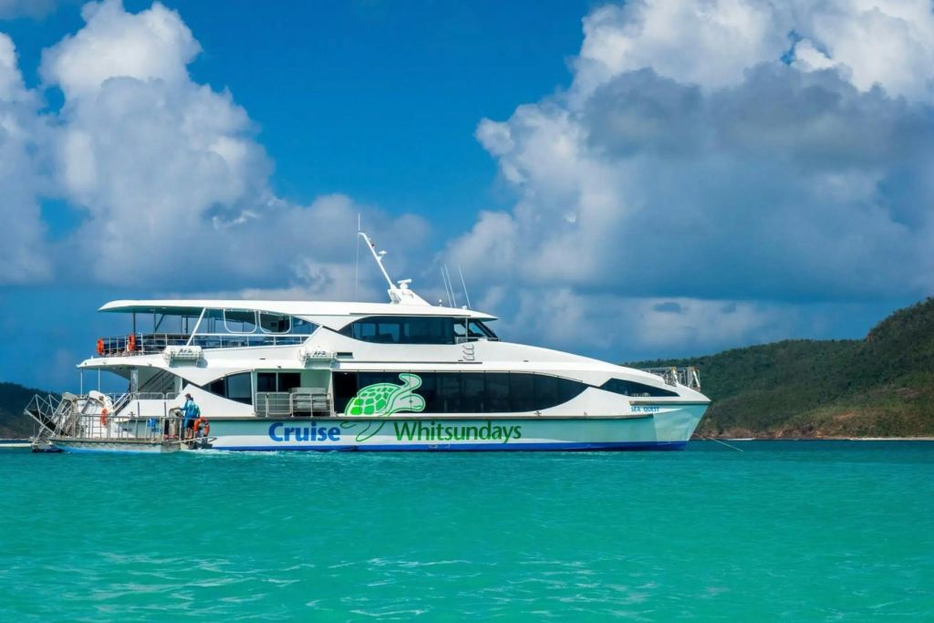 "Cruise Whitsundays" 와 거북이 로고가 있는 흰색 보트가 부분적으로 흐린 푸른 하늘 아래 청록색 물 위에 있습니다.