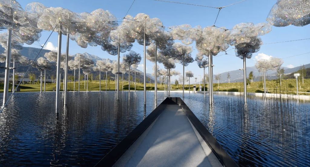 맑은 하늘 아래 산책로에서 바라본 물웅덩이 위의 반사 구름이 있는 금속성 나무 모양의 조각상.