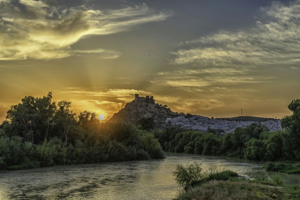 울창한 나무와 부분적으로 흐린 하늘로 둘러싸인 근처의 강이 흐르는 언덕 위 성 위로 석양을 감상해보세요.