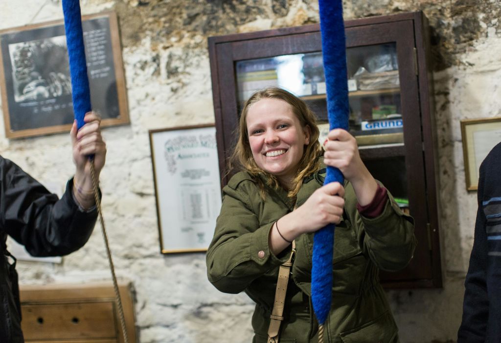 초록색 재킷을 입은 한 여성이 파란색 밧줄을 끌고 벽으로 둘러싸인 방 안으로 들어가는데, 그 뒤에는 액자에 들어간 서류와 나무 수납장이 있습니다.