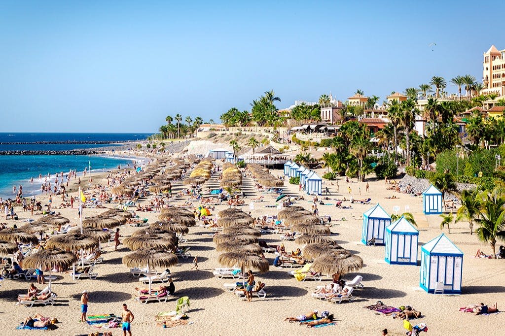 짚으로 지은 우산, 라운지 의자의 일광욕 의자, 카바나, 야자수와 건물을 배경으로 붐비는 해변.