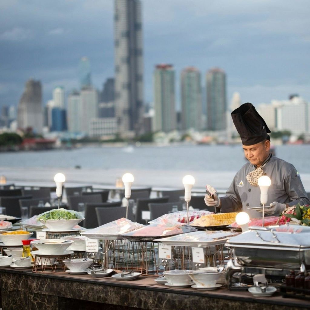 도시의 스카이라인과 강을 배경으로 야외 뷔페에서 키 큰 모자를 쓴 셰프가 음식을 준비합니다.
