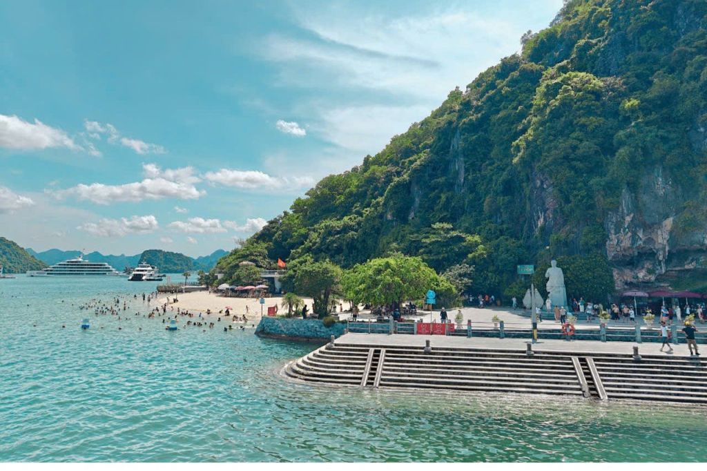 맑은 물, 울창한 푸른 언덕, 돌 계단, 동상, 수영하고 휴식을 취하는 사람들이 있는 경치 좋은 해변.