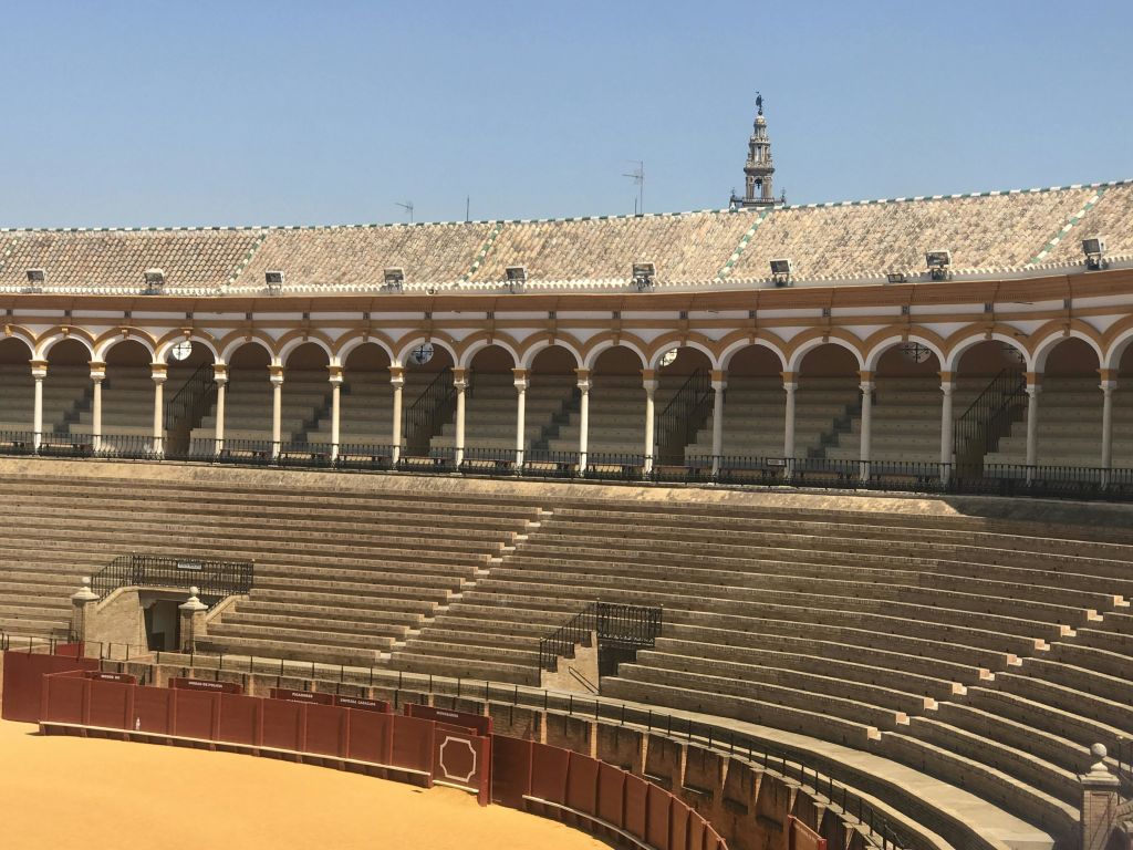 아치형 좌석 공간과 맑고 푸른 하늘 아래 배경으로 보이는 탑이 있는 텅 빈 역사적인 투우장의 모습입니다.
