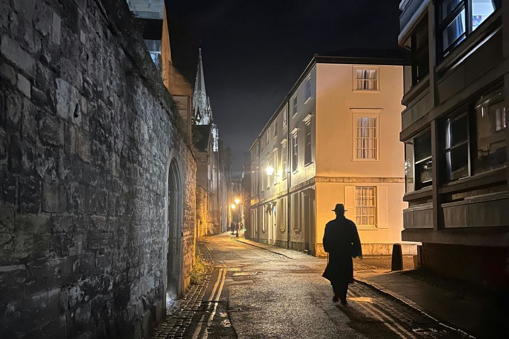 모자를 쓴 사람의 실루엣과 가로등 아래로 걸어 나가는 코트가 어둡게 빛나는 밤의 자갈길.