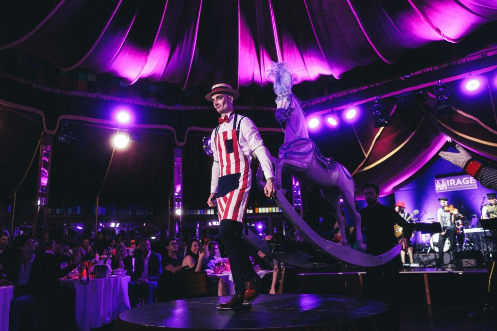 회전목마 옆 무대에서 빨간색과 흰색 줄무늬 의상과 밀짚 모자를 쓴 남성이 좌석에 앉은 관객을 배경으로 공연을 펼치고 있습니다.