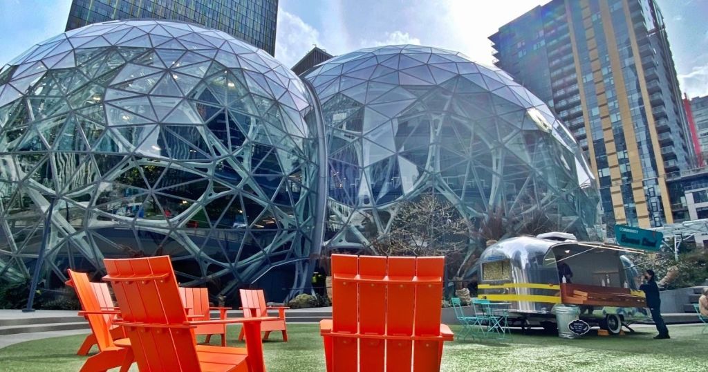 건물로 둘러싸인 기하학적 디자인의 두 개의 유리 돔 구조. 앞쪽에는 잔디 위에 주황색 의자가 놓여 있습니다.