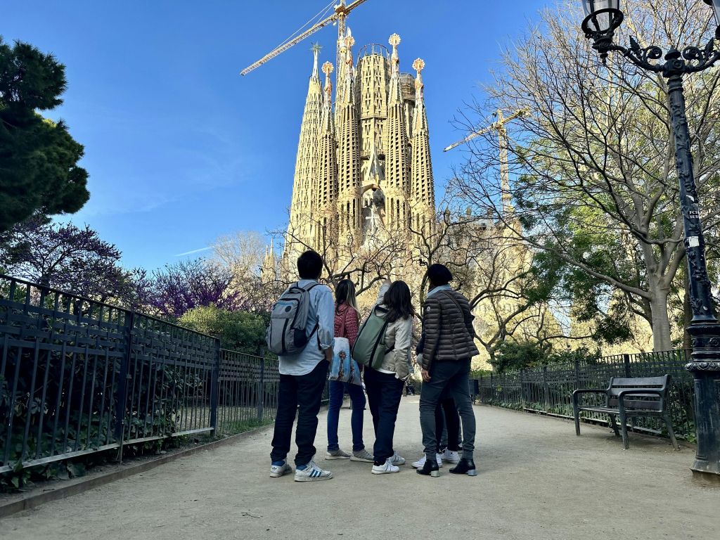 공원에 서 있는 다섯 명이 저 멀리 사그라다 파밀리아 성당을 바라보고 있습니다. 나무와 철제 울타리가 길에 늘어서 있습니다.