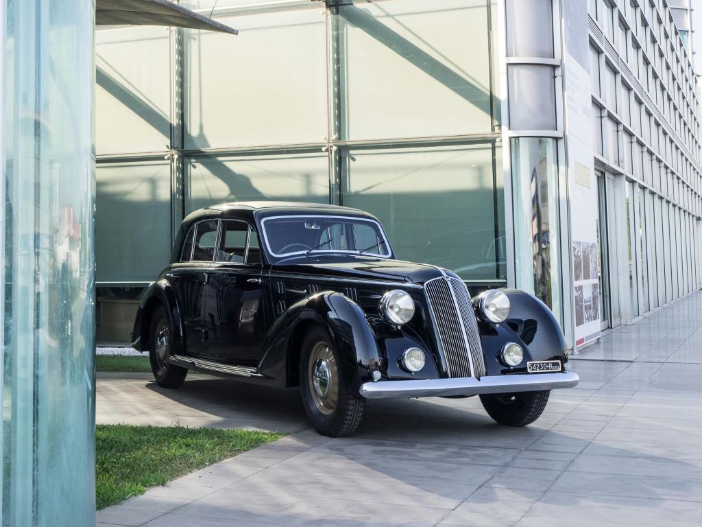 현대적인 유리 건물 근처의 타일 표면에 둥근 전조등이 달린 빈티지 검은색 자동차가 주차되어 있습니다.