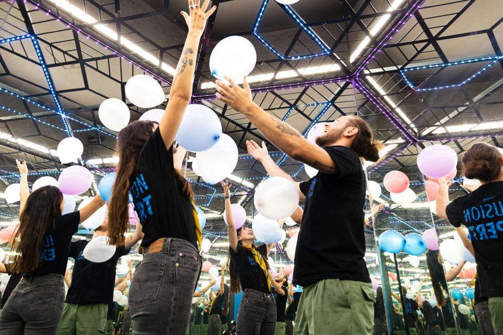 거울 벽과 화려한 조명이 있는 방에 있는 두 사람이 하얀 풍선을 공중에 던지며 장난스러운 분위기를 연출합니다.