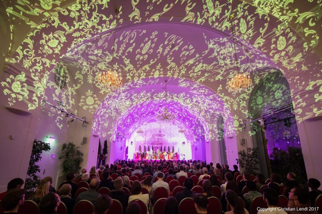 화려한 꽃무늬 조명과 샹들리에가 있는 호화로운 홀에서 관객이 무대 공연을 관람하고 있습니다.