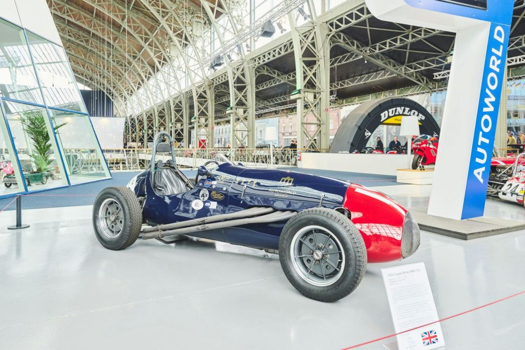 앞면이 빨간색이고 몸통이 파란색인 빈티지 레이스 카가 실내에 매끄러운 바닥에 전시되어 있습니다. 전시 플라크와 금속 트러스 구조가 보입니다.