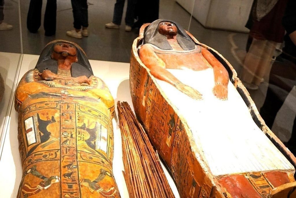 두 개의 고대 이집트 석관, 하나는 상형 문자로 장식되어 있고 다른 하나는 흰색 수의로 부분적으로 덮여 있으며 박물관에 전시되어 있습니다.