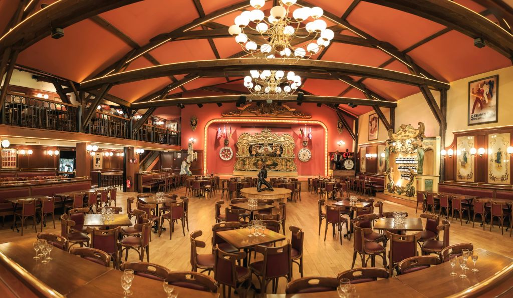 나무 테이블과 의자가 있는 크고 화려한 식당, 샹들리에, 정교한 장식이 있는 무대, 중이층.