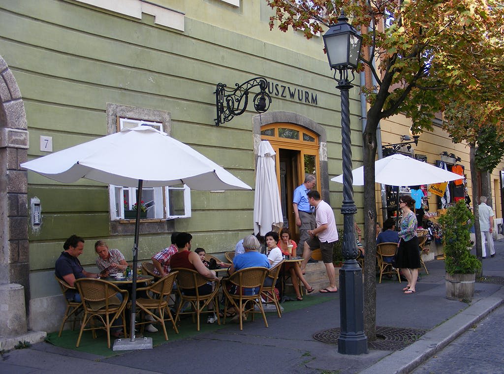 가로수가 늘어선 거리에 있는 커다란 흰색 우산 아래 "Ruszwurm" 간판이 있는 카페에 앉아 서 있는 사람들.