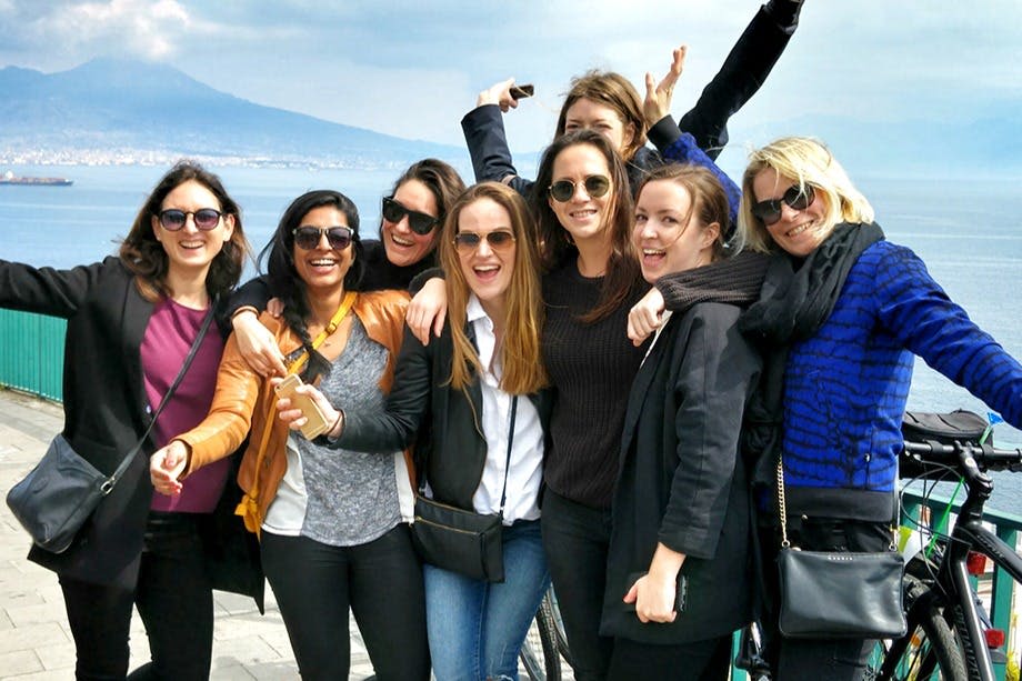 여덟 명의 여성이 산과 바다를 배경으로 해변 산책로에서 함께 웃으며 포즈를 취하고 있습니다.