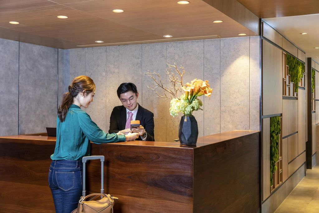 호텔 리셉션에 체크인한 한 여성이 리셉션 직원에게 전화를 건네고 있습니다. 그녀의 옆에는 여행 가방이 놓여 있고, 카운터에는 꽃이 장식되어 있습니다.
