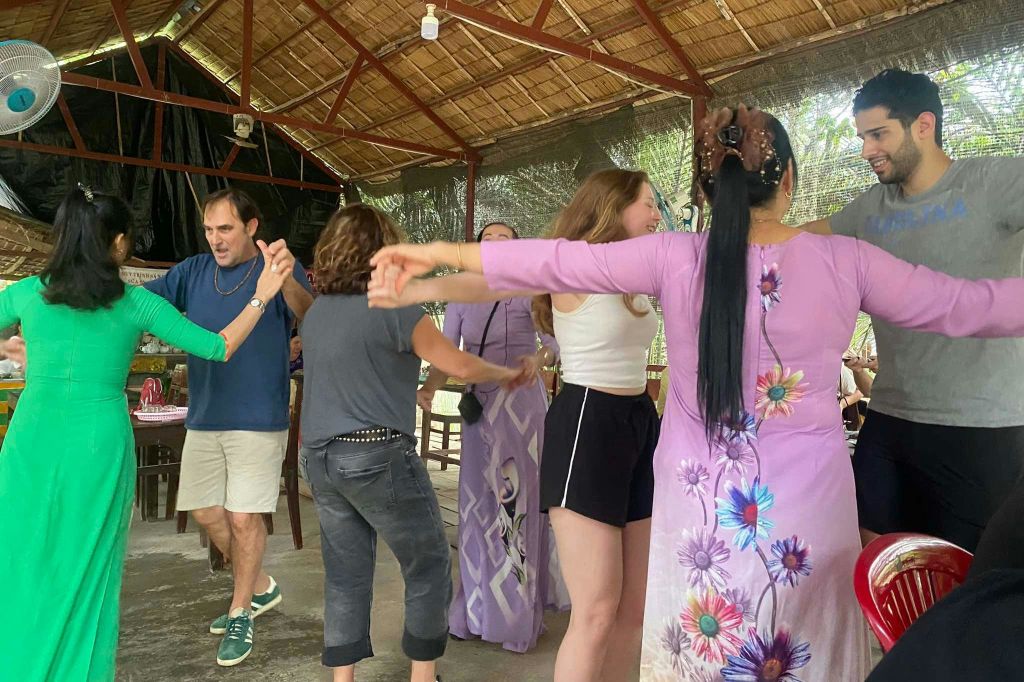 초가 지붕 아래에서 원을 그리며 미소를 짓고 있는 사람들. 한 사람이 꽃무늬가 있는 보라색 드레스를 입습니다.