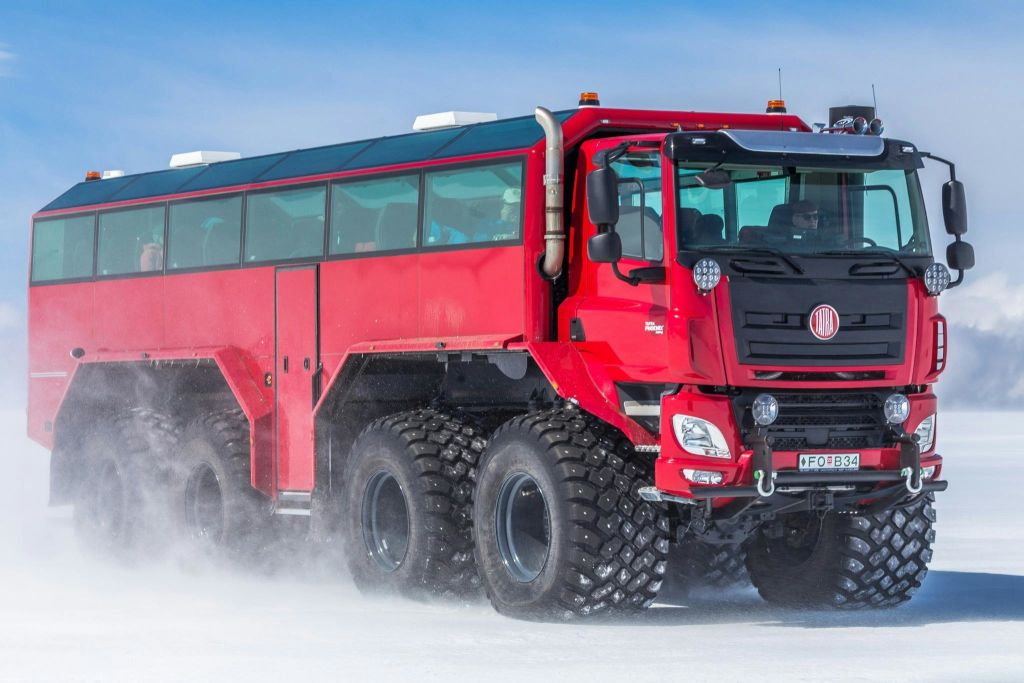 여러 개의 타이어가 달린 커다란 빨간색 오프로드 차량이 맑고 푸른 하늘 아래 눈 덮인 지형을 주행하고 있습니다.