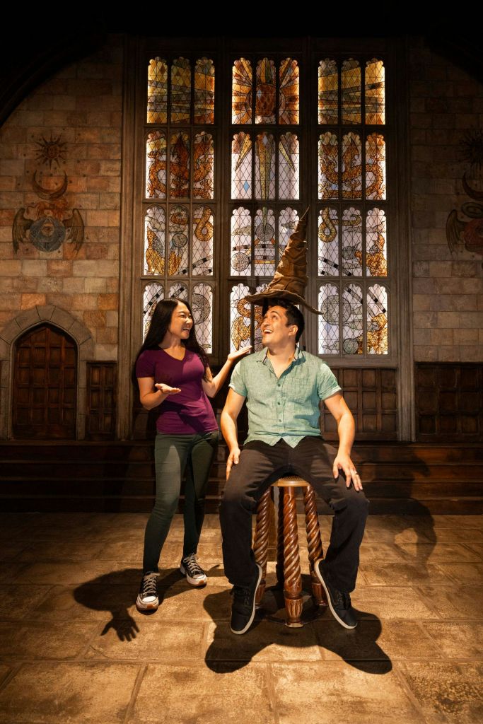 돌방에 있는 두 사람. 하나는 마법사 모자를 쓰고 화려한 스테인드글라스 창문 앞에 앉은 채로 서 있습니다.