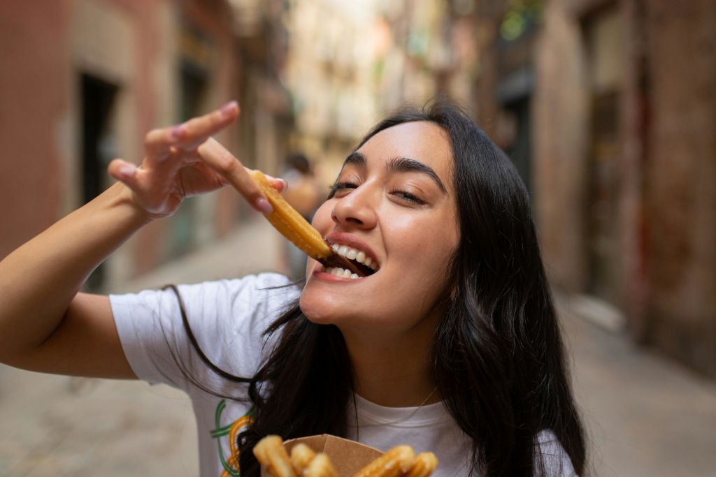 어두운 머리카락이 긴 여성이 좁은 골목에서 츄로를 먹으며 더 많은 츄로가 담긴 용기를 들고 미소를 짓고 있습니다.