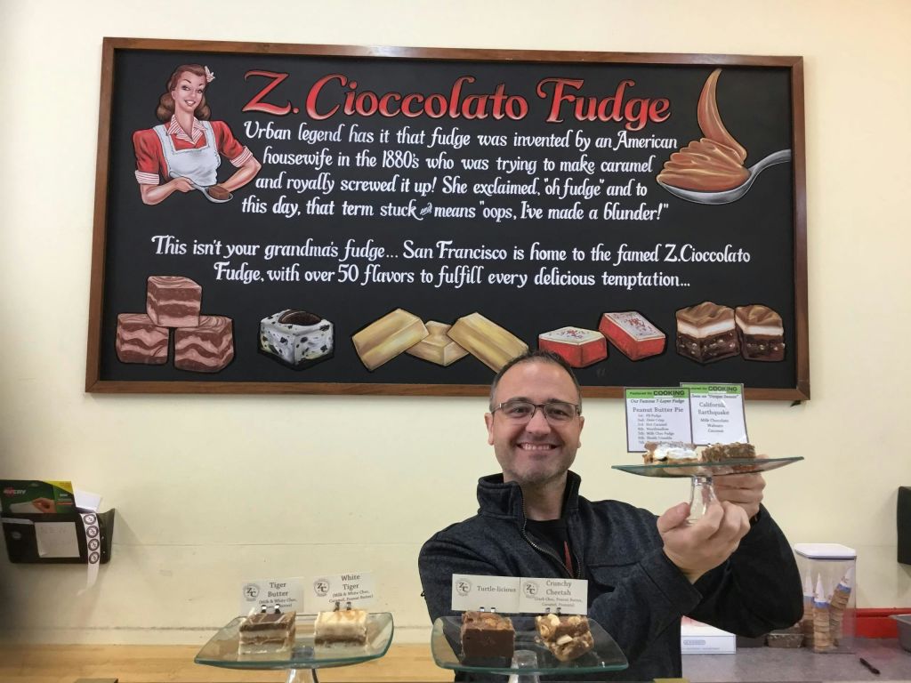한 남성이 'Z. Cioccolato Fudge' 벽면 사인 앞에 디저트 접시를 들고 퍼지 샘플을 전시하며 미소를 짓고 있습니다.