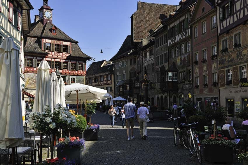 유서 깊은 건물이 있는 자갈길을 걷는 사람들, 우산, 화분, 자전거가 놓인 야외 카페.