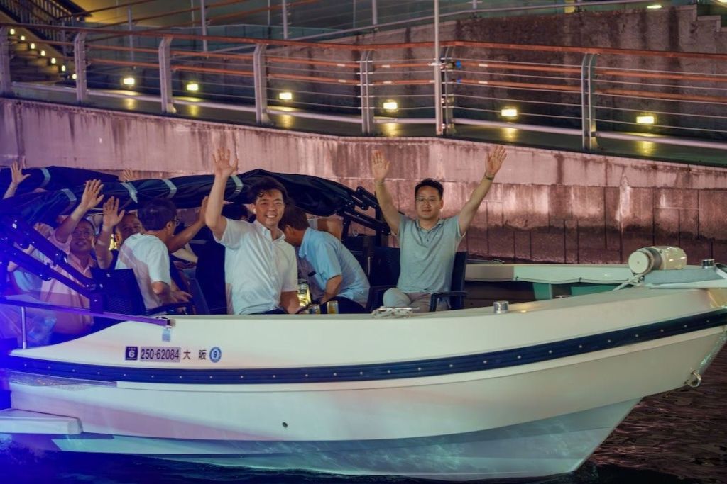 보트를 탄 두 남자가 카메라를 향해 기분 좋게 손을 흔들고 있으며, 배경에 세 번째 남자가 무언가를 조정하고 있습니다. 그 뒤로 불이 켜진 통로가 있습니다.