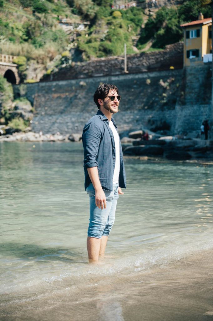 돌담과 노란색 건물 근처의 얕은 물에 서서 선글라스와 캐주얼한 옷을 입고 녹지를 배경으로 서 있는 남성.