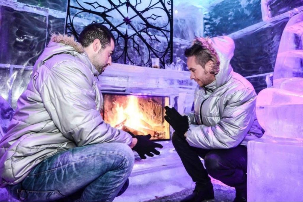 후드가 달린 재킷을 입은 두 사람이 푸른 조명과 얼음 장식으로 얼음 환경의 벽난로 근처에서 손을 따뜻하게 합니다.