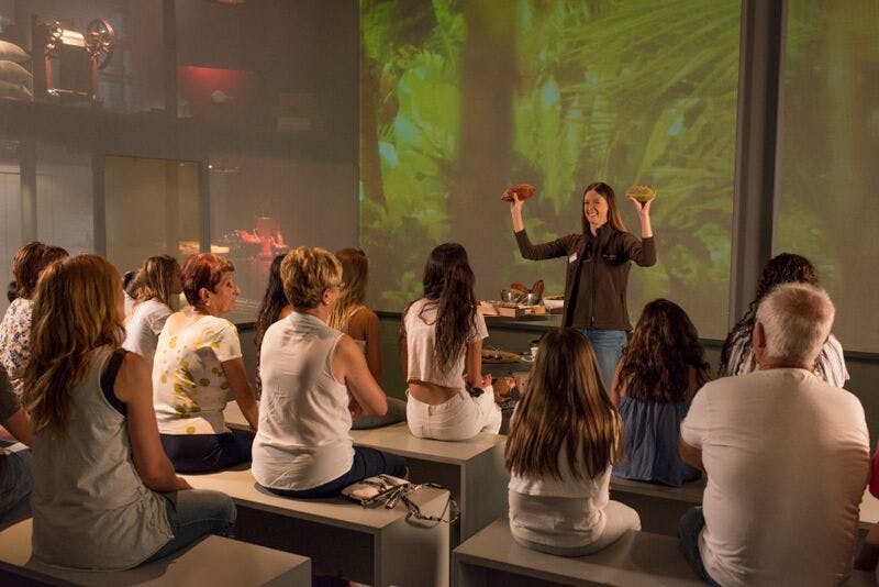 한 여성이 배경이 투영된 방에서 앉은 청중과 대화하는 동안 두 개의 물건을 들고 있습니다.