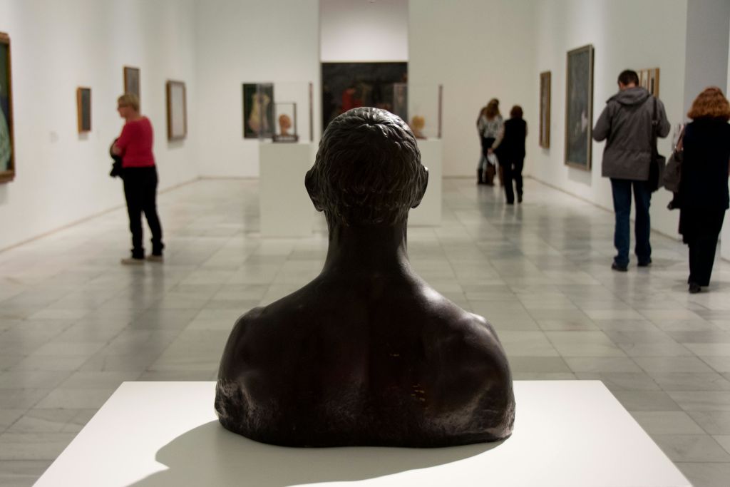 전경에 청동 흉상이 있고 벽과 받침대를 배경으로 사람들이 예술 작품을 감상하는 갤러리입니다.