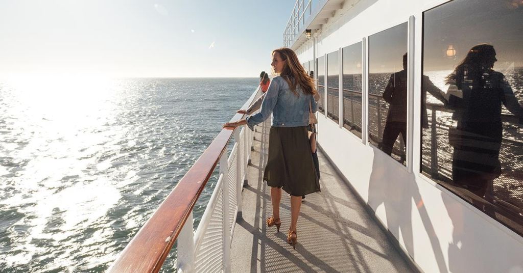 맑고 화창한 하늘 아래 오른쪽에 바다가 보이는 배의 갑판을 걷고 있는 데님 재킷을 입은 여성.