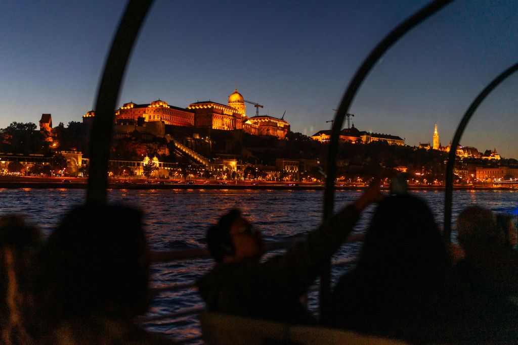 물 건너 언덕에 조명이 켜진 역사적인 건물의 전망을 감상하며 밤에 보트를 타는 사람들.