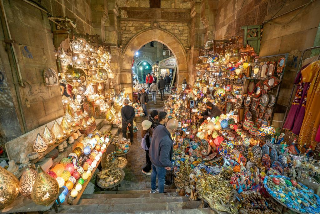 화려한 램프와 장식품으로 가득한 번화한 시장 노점에는 사람들이 둘러보고 있습니다. 아치형 통로를 배경으로 더 많은 상점이 나옵니다.