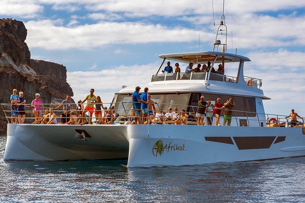 바위 절벽과 부분적으로 흐린 하늘을 배경으로 사람들이 탑승하여 물가에서 화창한 하루를 즐기는 대형 쌍동선입니다.