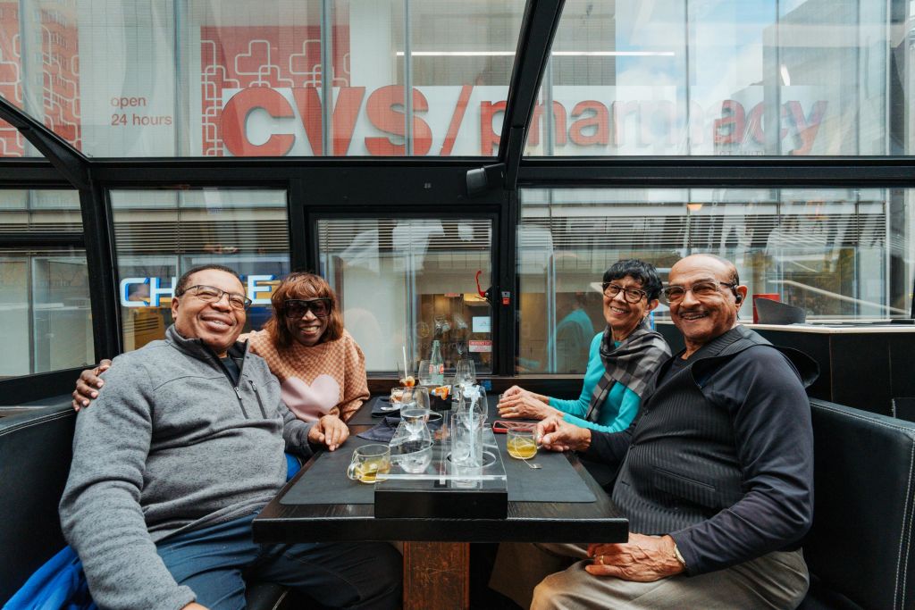 세 사람이 테이블에 앉아 음료와 빈 잔을 들고 미소를 짓고 있습니다. 배경에는 "약국" 이라는 단어가 있는 유리 벽이 있습니다.