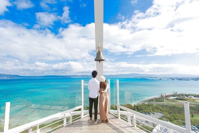 청록색 바다가 내려다보이는 나무 플랫폼에 서 있는 커플로, 커다란 종소리와 밝고 푸른 하늘이 배경으로 펼쳐집니다.