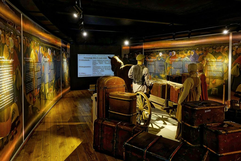 카트와 수하물이 있는 기간 의류의 마네킹이 전시되어 있습니다. 벽은 어두운 조명 아래에서 역사적인 텍스트와 이미지를 표시합니다.