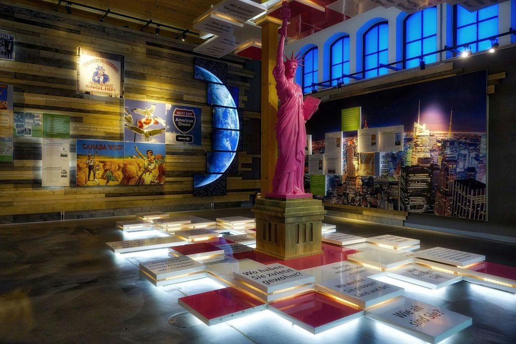조명 디스플레이 패널과 다양한 벽 포스터로 둘러싸인 분홍색 자유의 여신상 복제품이 있는 실내 전시품.