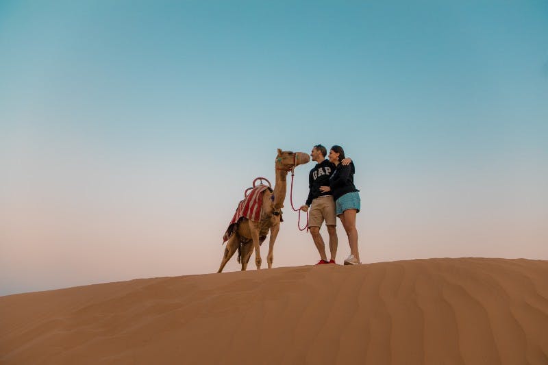 두 사람과 낙타 한 마리가 맑은 하늘 아래 모래 언덕에 서 있고, 낙타는 빨간색과 흰색 줄무늬로 장식되어 있습니다.